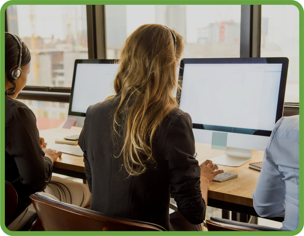 People wearing headphones type on desktop computers in an bright open plan office. They sit side by side at a desk facing a large window.